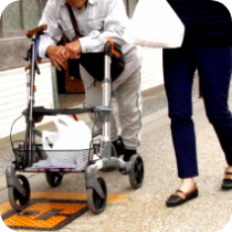 手押し車を押して歩行する高齢者と、付き添うスタッフの写真