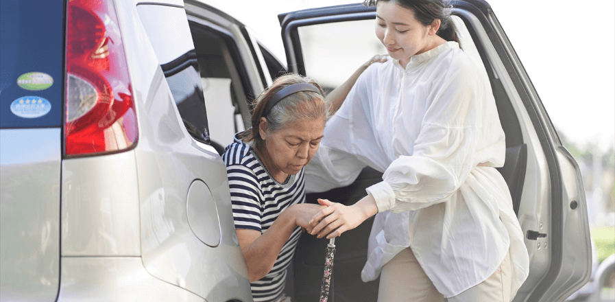 杖を持った高齢女性が車から降りるのを手助けする女性スタッフの写真